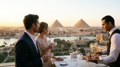 صورة واقعية لزوجين يستمتعان بوقت الغروب على شرفة فندق فاخر في القاهرة، مع إطلالة بانورامية ساحرة على أهرامات الجيزة ونهر النيل، بينما يقدم لهما نادل صينية حلويات