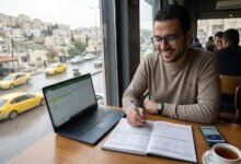 شاب أردني مبتسم يقوم بتنظيم الميزانية الشخصية وتخطيط المصروفات باستخدام لابتوب يعرض جدول بيانات ودفتر ملاحظات، وذلك أثناء جلوسه في مقهى يطل على شارع ممطر وسيارات أجرة صفراء في مدينة عمان، الأردن.